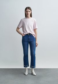 Nő világos rózsaszín mintás pólóban, kék virágdíszes, rövidített farmernadrágban, fehér zokniban és fehér hegyes orrú magas sarkú cipőben, egyszerű háttér előtt állva.