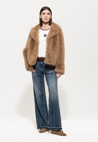 Braune Kunstfelljacke mit großen Knöpfen, kombiniert mit einem weißen Hemd mit rotem Stickerei, weit geschnittenen Denim-Jeans und beigen Schuhen.