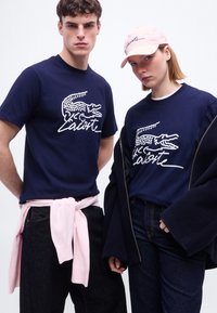 Νεαρός άνδρας και γυναίκα που φορούν μπλε σκούρα πουκάμισα Lacoste με λευκό λογότυπο κροκόδειλου· η γυναίκα φοράει επίσης ροζ καπέλο Lacoste και σκούρο μπουφάν.