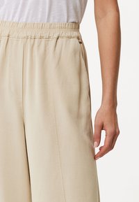 Pantalons larges beige avec taille élastique, fabriqués en tissu doux et lisse. Comprend des poches latérales et une coupe décontractée pour plus de confort.