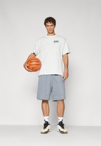 Lys grå bomulds T-shirt med lille flerfarvet "Nike" logo, parret med blå shorts. Holder en orange basketball. Bærer sorte og hvide sneakers.