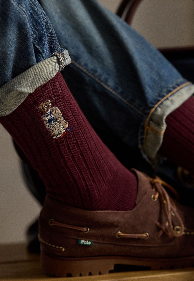 Meias caneladas cor bordô com um logótipo bordado de um urso a usar um uniforme azul, combinadas com sapatos de camurça castanhos. Jeans de ganga com a bainha dobrada.