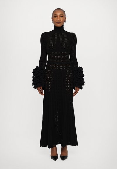 Robe noire en tricot à manches longues, avec un col montant et des poignets à volants. Le design comprend un motif de trous sur l'ensemble du tissu.