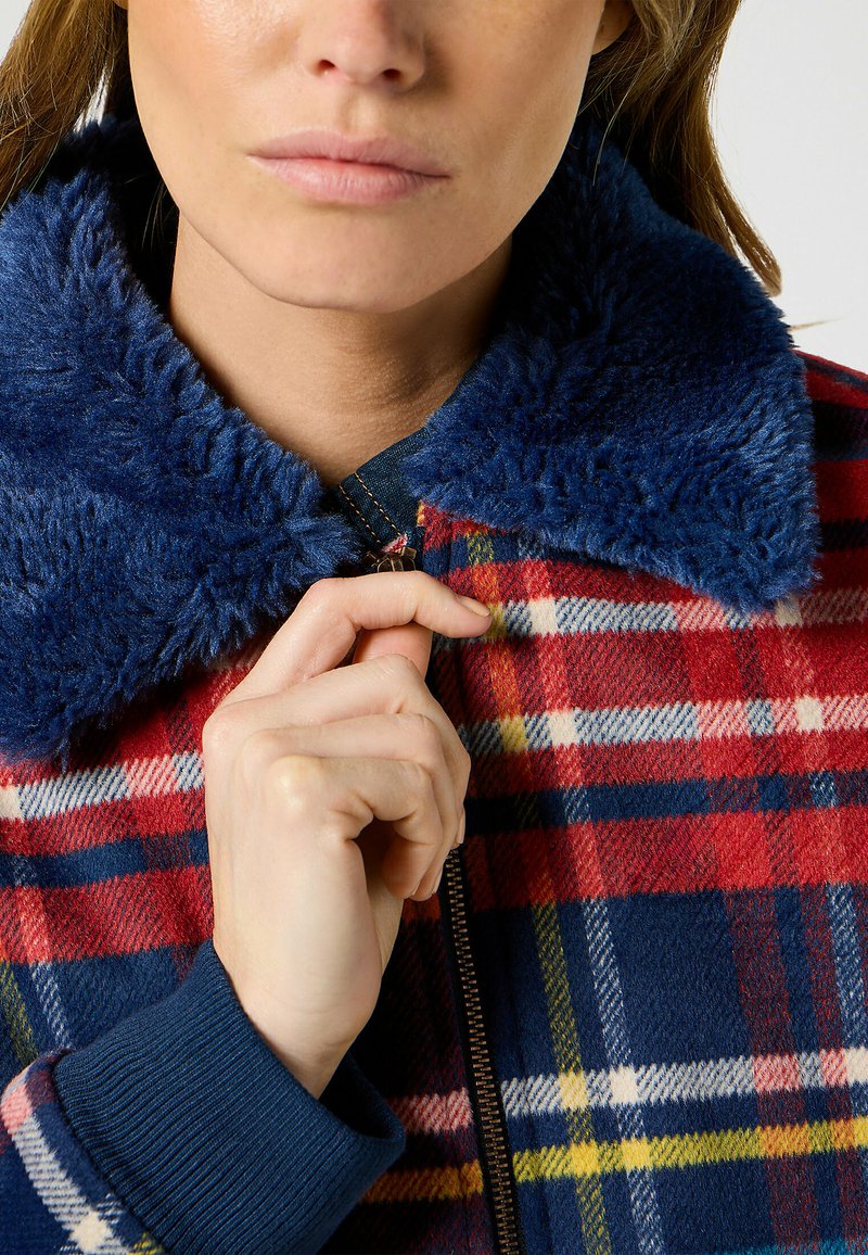 Giacca a quadri con un motivo rosso, blu e giallo, dotata di un colletto blu morbido e polsini blu a coste. Dettaglio con cerniera visibile.