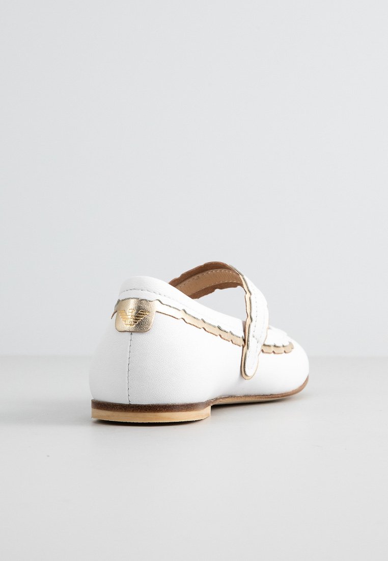 Scarpa Mary Jane in pelle bianca con rifiniture e cinturino dorato, suola piatta in legno, vista da dietro e di lato su una superficie e sfondo grigi.