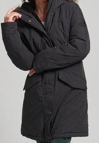 Musta eristetty parka, jossa on karvakauluri, vetoketjullinen etuosa, isot etutaskut ja lämpöä lisäävä teksturoitu kangas.