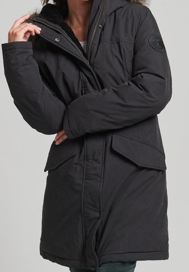 Musta eristetty parka, jossa on karvakauluri, vetoketjullinen etuosa, isot etutaskut ja lämpöä lisäävä teksturoitu kangas.