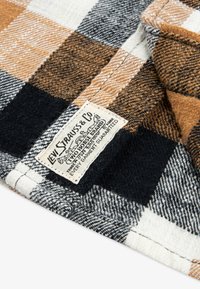 Camicia in flanella con un motivo a quadri nero, bianco e arancione. Ha un'etichetta in tessuto cucita di lato con i dettagli del marchio.