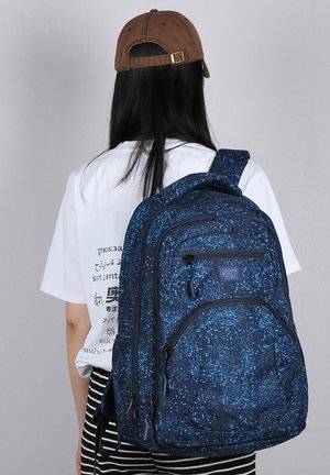 Blue patterned backpack with a textured fabric, multiple zippered compartments, and adjustable straps, worn on the back.