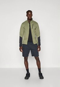 Veste zippée vert olive avec manches noires, portée sur un T-shirt noir ; associée à un short foncé et des baskets noires. Design simple, style fonctionnel.
