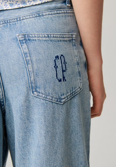 Tasca posteriore di jeans denim azzurri chiari con logo "CP" ricamato blu scuro indossati da una persona con un top bianco e blu a fiori.