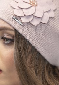 Bonnet en maille rose clair avec une décoration de fleur en feutre superposée et une étiquette de marque en argent. Texture lisse et ajustement serré.