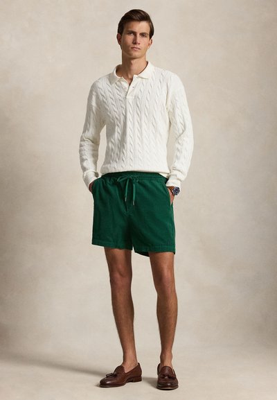 Homme portant un pull en maille torsadée blanc, un short vert à cordon, des mocassins marron et une montre-bracelet, debout les mains dans les poches.