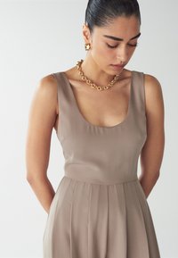 Beige mekko ilman hihoja, jossa on pyöreä pääntie, taitettu malli ja sileä rakenne. Yhdistetty massiiviseen kultaketjuun.