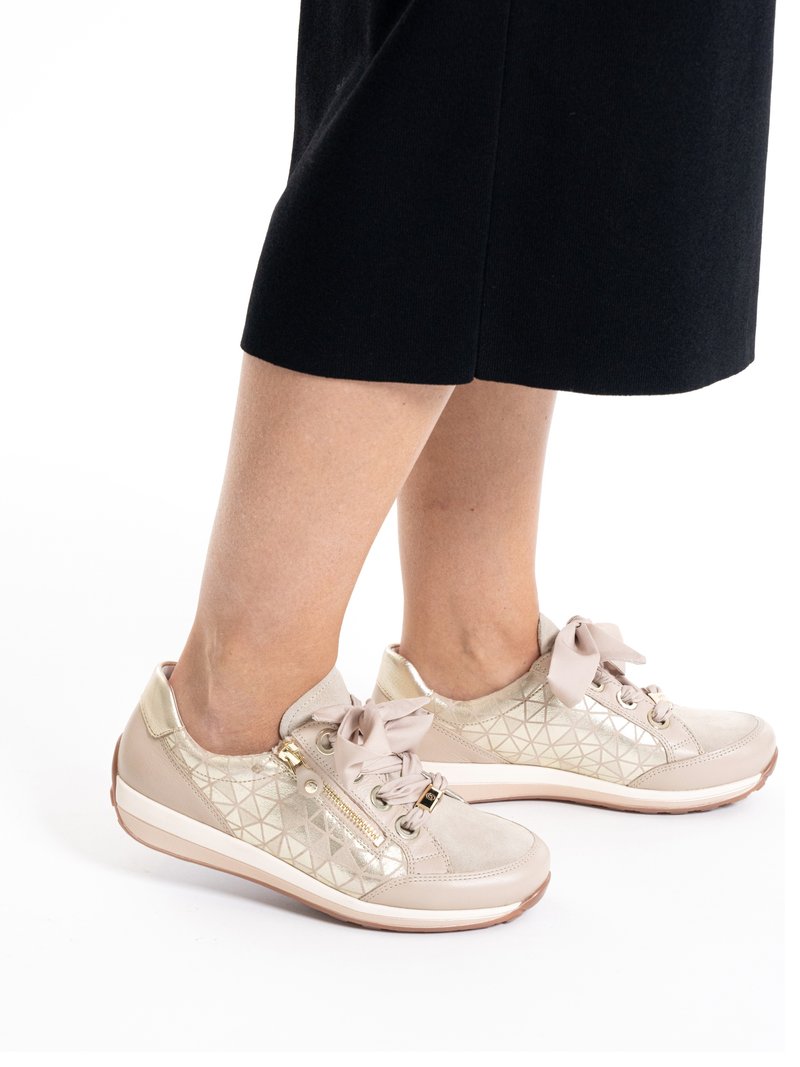 Femme portant des baskets beiges avec des motifs géométriques, des lacets ruban et des fermetures éclair latérales, associées à un pantalon noir raccourci.