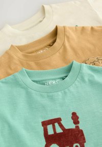 Trois t-shirts à col rond : crème, beige et turquoise clair. Le t-shirt turquoise arbore un graphique de train brun ; tous ont des cols côtelés et un tissu texturé.