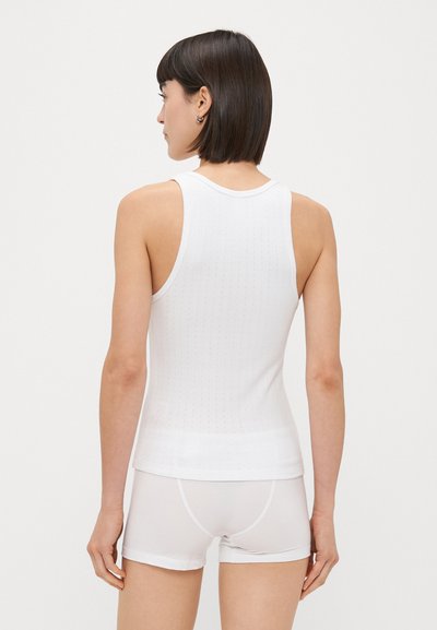 Balta bez piedurknēm auduma virsotne ar teksturētu apdari, pieguļoša silueta un apaļu kakla izgriezumu, kombinēta ar baltām šortiem.