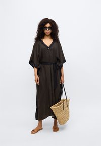 Keftan à motifs noirs avec un col en V et des manches larges, cintré à la taille avec un lien. Porté avec un sac cabas en paille et des sandales.