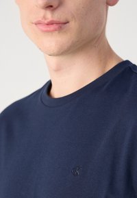 Tamsiai mėlynas medvilninis marškinėliai su apvaliu kaklu ir trumpomis rankovėmis, kuriuose dešinėje krūtinės pusėje yra mažas siuvinėti 'ck' logotipas.