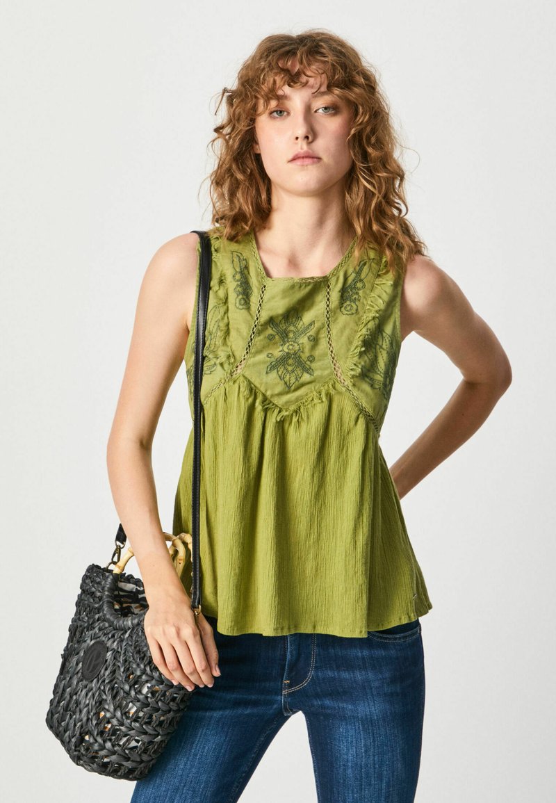 Top verde senza maniche con design floreale ricamato e texture plissettata, abbinato a jeans blu scuri e una borsa nera intrecciata.