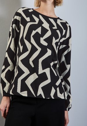 Kobieta ubrana w czarną bluzkę z długim rękawem z odważnymi, abstrakcyjnymi, białymi zygzakowatymi wzorami i czarne spodnie na jednolitym szarym tle.