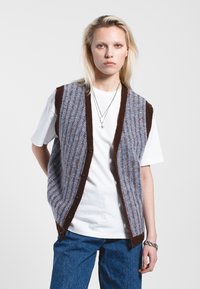 Gilet en tricot présentant un motif zigzag bleu et marron, avec une bordure et des boutons marron, porté sur un t-shirt blanc et associé à un jean bleu.