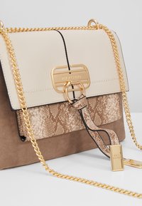 Bolso de mano beige y marrón con herrajes dorados, que presenta una sección superior lisa, un medio con estampado de serpiente texturizado y un fondo de ante suave. Incluye una cadena como strap.