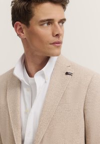 Beiger strukturierter Blazer mit Stehkragen über einem weißen Hemd mit Knöpfen. Verfügt über ein kontrastierendes gestreiftes Detail am Revers.