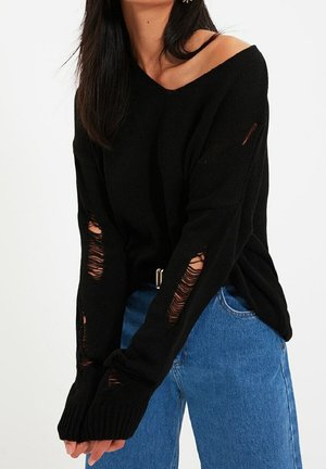 Femme portant un pull noir à épaules dénudées avec plusieurs trous usés et un jean bleu taille haute, sur un fond uni.