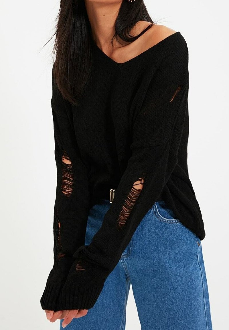 Femme portant un pull noir à épaules dénudées avec plusieurs trous usés et un jean bleu taille haute, sur un fond uni.