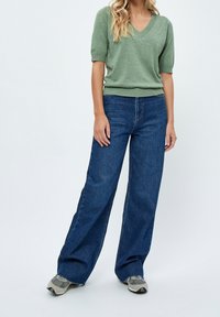 Zielony sweter z krótkim rękawem i dekoltem w serek połączony z niebieskimi dżinsami o wysokim stanie i szerokimi nogawkami oraz szarymi sneakersami. Tkanina wydaje się być miękka i ma luźny krój.