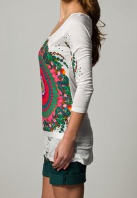 Vit långärmad topp med ett färgglatt cirkulärt mönster i grönt, rosa och blått, med rund halsringning, tillsammans med mörkgröna shorts.