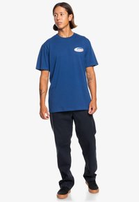 Navy blue cotton t-shirt with a round neck and short sleeves, featuring a small multi-colored logo on the chest, paired with dark pants.