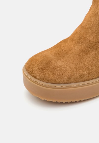 Tan suede ankle boot with a rounded toe, thick rubber sole, and a smooth texture; features a stitched seam along the sole edge.