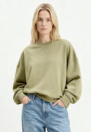 Donna con capelli biondi che indossa una felpa oversize verde oliva e jeans a vita alta azzurri, mani nelle tasche, sfondo neutro.