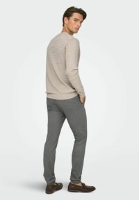 Beežikas tekstuuriga kampsun, hallid kitsad püksid ja pruunid mokassiinid. Modell seisab seljaga vaataja poole, käed taskutes, näidates riietuse lõiget.