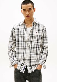White and black plaid shirt with long sleeves, button-down collar, and logo detail. Worn with a white undershirt and dark denim jeans.