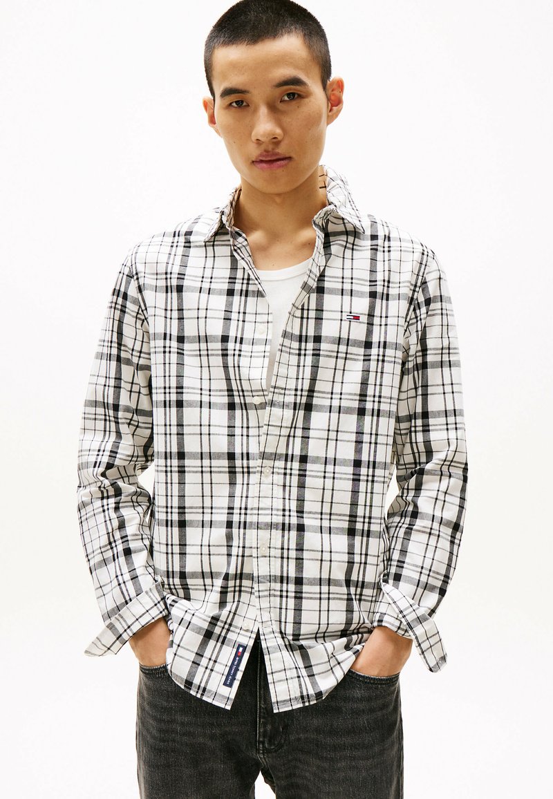 White and black plaid shirt with long sleeves, button-down collar, and logo detail. Worn with a white undershirt and dark denim jeans.