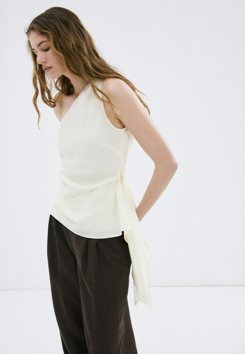 Femme aux longs cheveux portant un haut blanc sans manches asymétrique et un pantalon noir ample, debout devant un fond blanc uni.