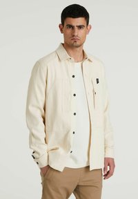 Camicia button-up beige chiaro con colletto, due tasche frontali e bottoni neri. Indossata sopra un maglione bianco a trama.