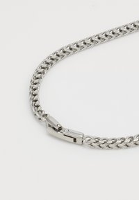 Collier en métal argenté avec de grosses maillons entrelacés et un fermoir rectangulaire gravé "GUESS" sur un fond uni.