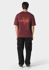 Henkilö, jolla on yllään viininpunainen T-paita, jossa on oranssi Pegasos ja teksti "Pas De Monaco" selässä, mustat cargo-housut ja mustat lenkkarit.