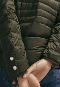 Veste matelassée vert foncé avec motif en losanges, dotée d'un détail de manchette boutonnée et d'une texture lisse. La manche est légèrement retroussée.