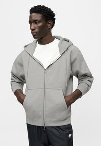 Γκρι φούτερ με φερμουάρ, ευρύχωρη κουκούλα, ριμπ μανσέτες και μπροστινές τσέπες, φορεμένο πάνω από λευκό μπλουζάκι. Λείος υφής, καθημερινό σχέδιο.