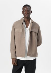 Tan, soft fabric jacket with two chest pockets, snap closure, and a classic collar, worn over a white shirt and black jeans.