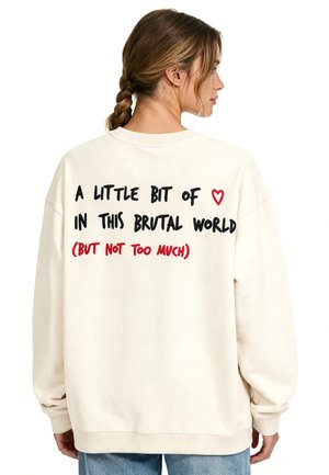 Donna con capelli intrecciati che indossa una felpa beige con un testo sulla schiena che dice "A little bit of heart in this brutal world (but not too much)"