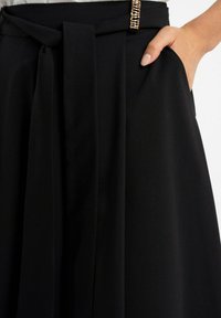 Czarna spódnica z miękkiego materiału, z wiązanym detalem w talii, bocznymi kieszeniami oraz dekoracyjnym metalowym akcentem w pasie.