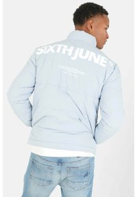 Giacca puffer azzurra chiara con grande testo bianco "SIXTH JUNE" sul retro, caratterizzata da un design trapuntato e colletto rialzato. Indossata sopra jeans di denim.