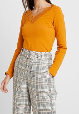 Vrouw draagt een mosterdkleurige top met lange mouwen en kanten V-hals en hooggetailleerde geruite broek met een gestrikte ceintuur, handen deels in de zakken.