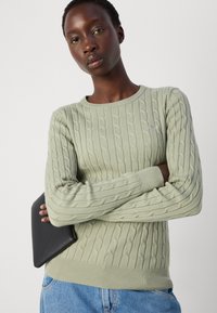 Maglione verde chiaro a maglia con collo rotondo e orlo a coste. Presenta un motivo testurizzato ed è abbinato a una pochette nera.